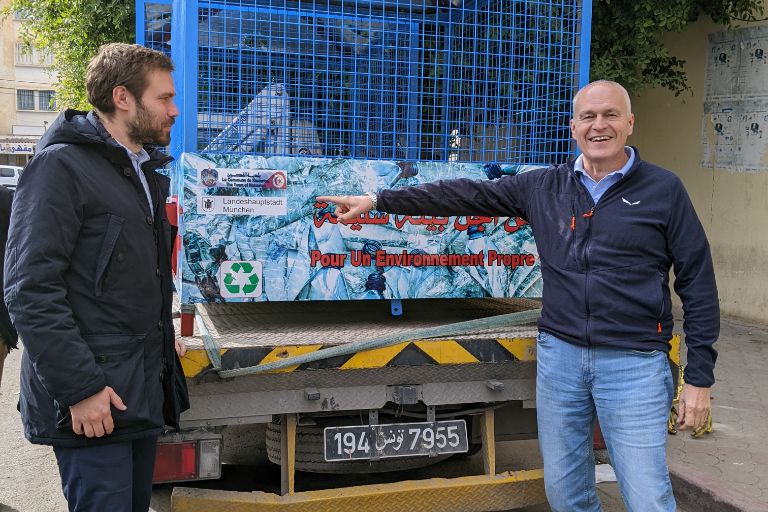 Zwei Personen stehen neben einem Recycling-Container mit den Logos der Partner-Kommunen