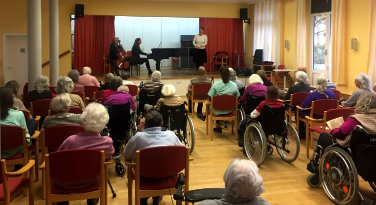 Senior*innen sitzen in einem Sall und hören der Klavier-Musik zu.