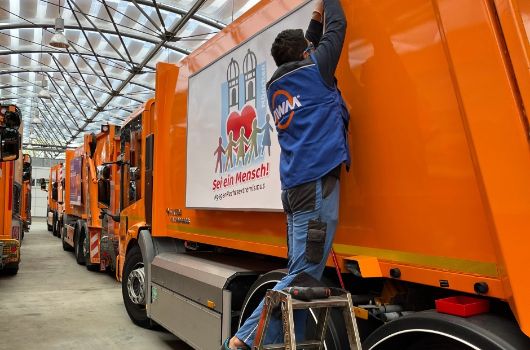 Mitarbeiter des Abfallwirtschaftsbetriebs München bringt ein Seitenschild mit dem Logo der Kampagne "Sei ein Mensch | #gegenRechtsextremismus" an einem Müllfahrzeug an.