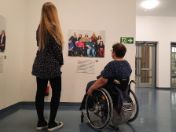 Foto: Von hinten sind zwei Frauen zu sehen. Sie schauen sich ein großes Foto an. Das Foto hängt an einer Wand in einem Flur. Eine Frau steht. Die andere Frau sitzt im Rollstuhl.