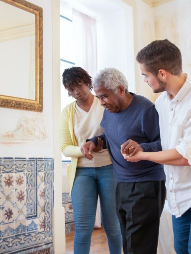 Zwei Jüngere stützen einen Älternen beim gehen. 